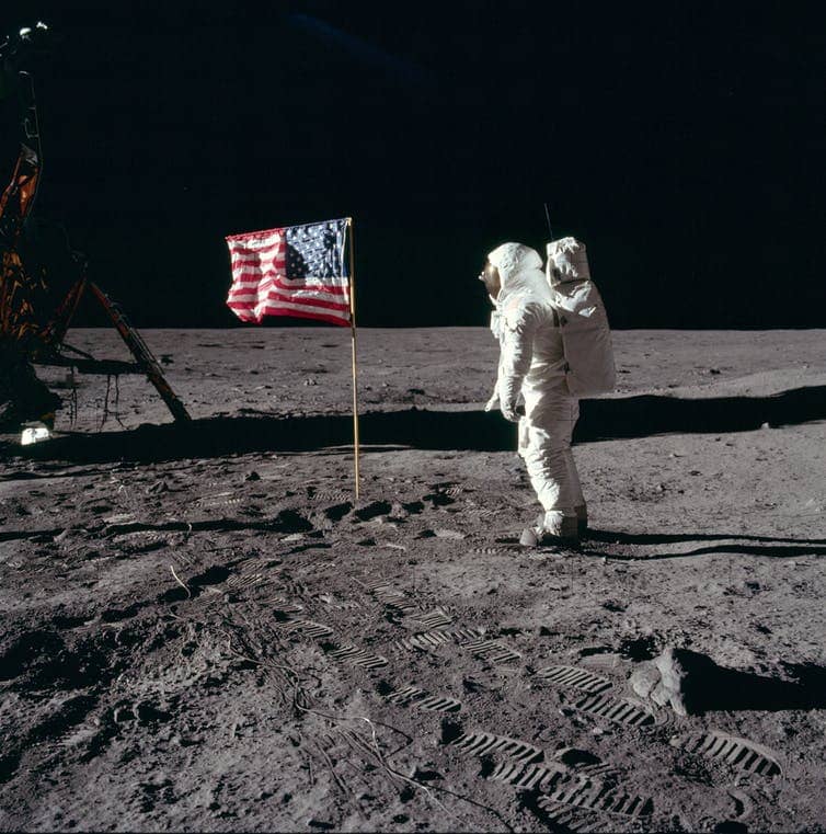 Buzz Aldrin in a spacesuit on the surface of the Moon next to the U.S. flag.